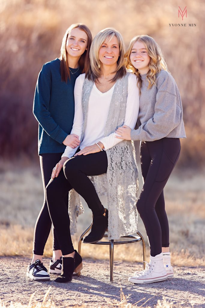 A mom and two teenage daughters sit on a stool in a field in Thornton, Colorado during their family photoshoot.