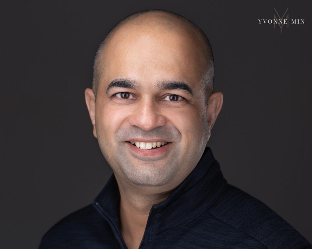 Headshot on a dark gray backdrop of a professional man in a black sweater by Yvonne Min Photography.