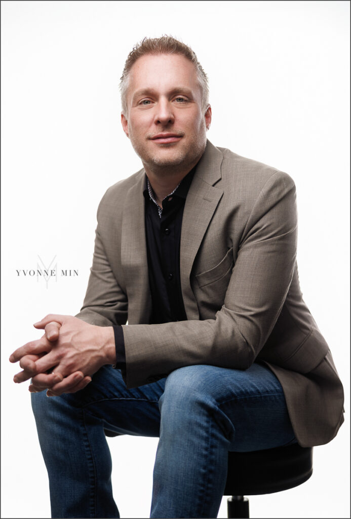 A photograph of a man sitting on a stool photographed by Yvonne Min Photography on a white backdrop at her studio in Thornton, Colorado.