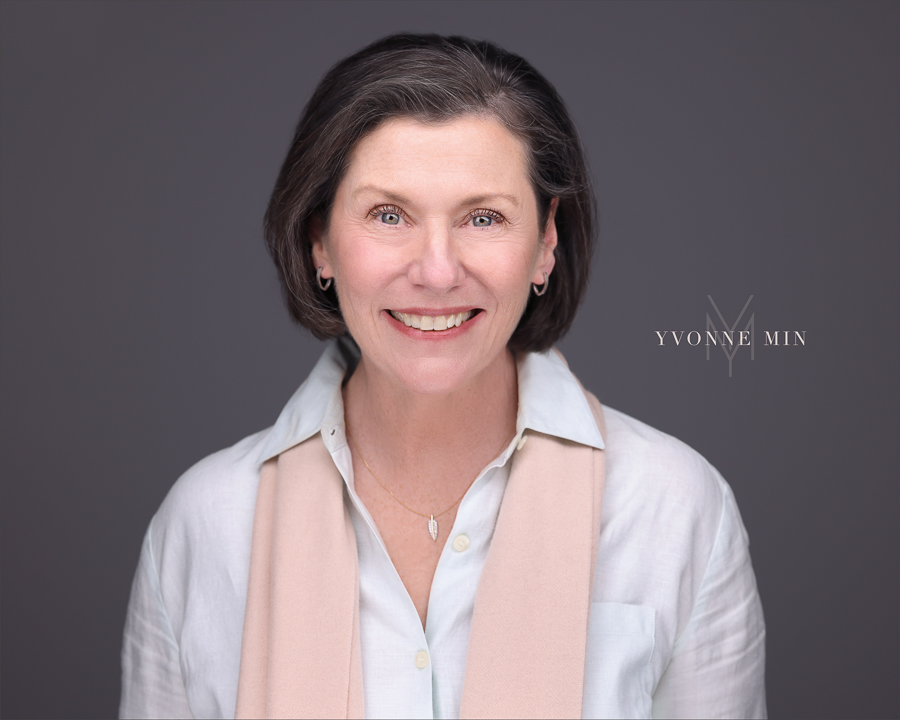 A headshot of a professional woman in a white shirt photographed on a dark gray backdrop in the studio of Yvonne Min Photography in Thornton, Colorado.