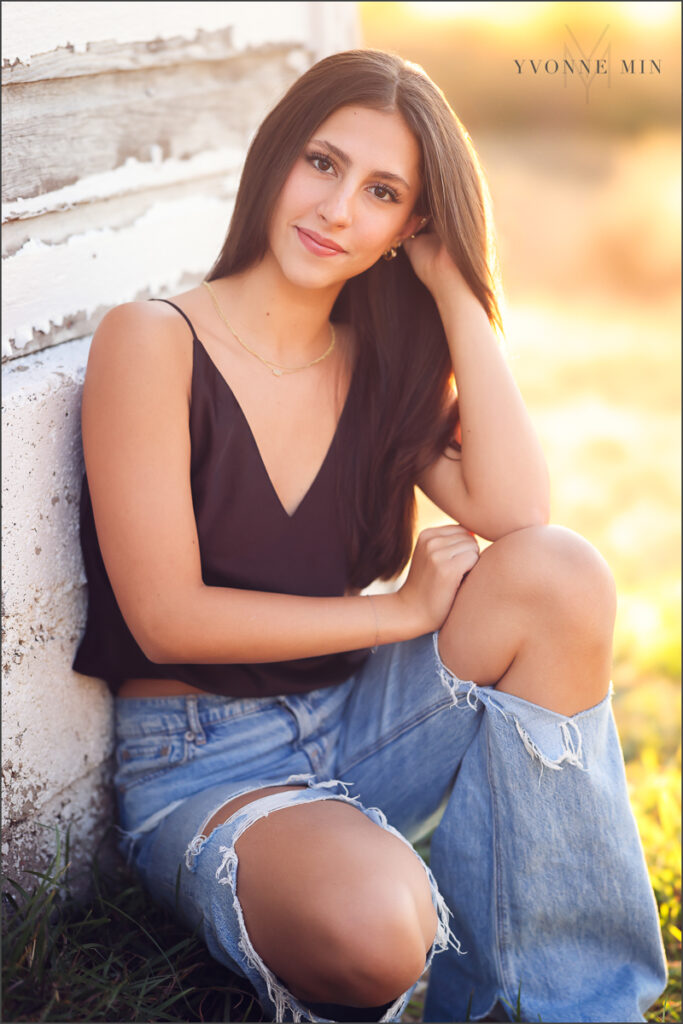 A young teen girl squats against a white barn wall during her family photography session with Yvonne Min Photography in Thornton, Colorado.