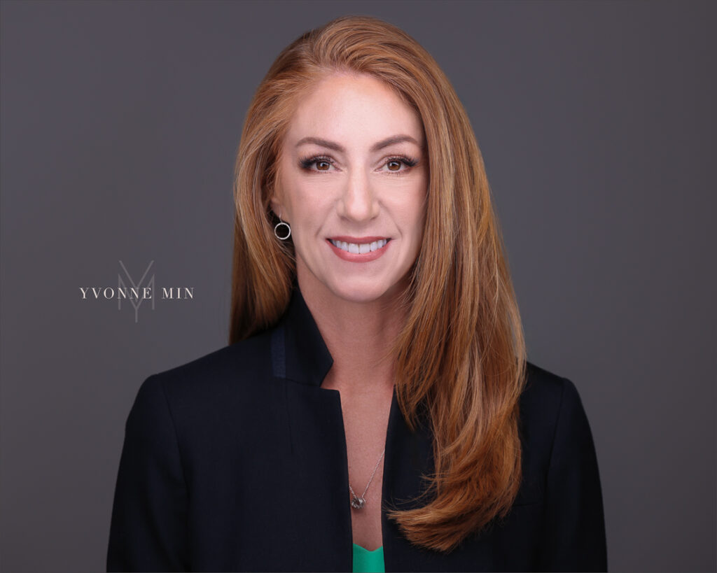 A corporate headshot of a professional woman with red hair photographed on a dark gray backdrop at the studio of Yvonne Min Photography in Thornton, Colorado.