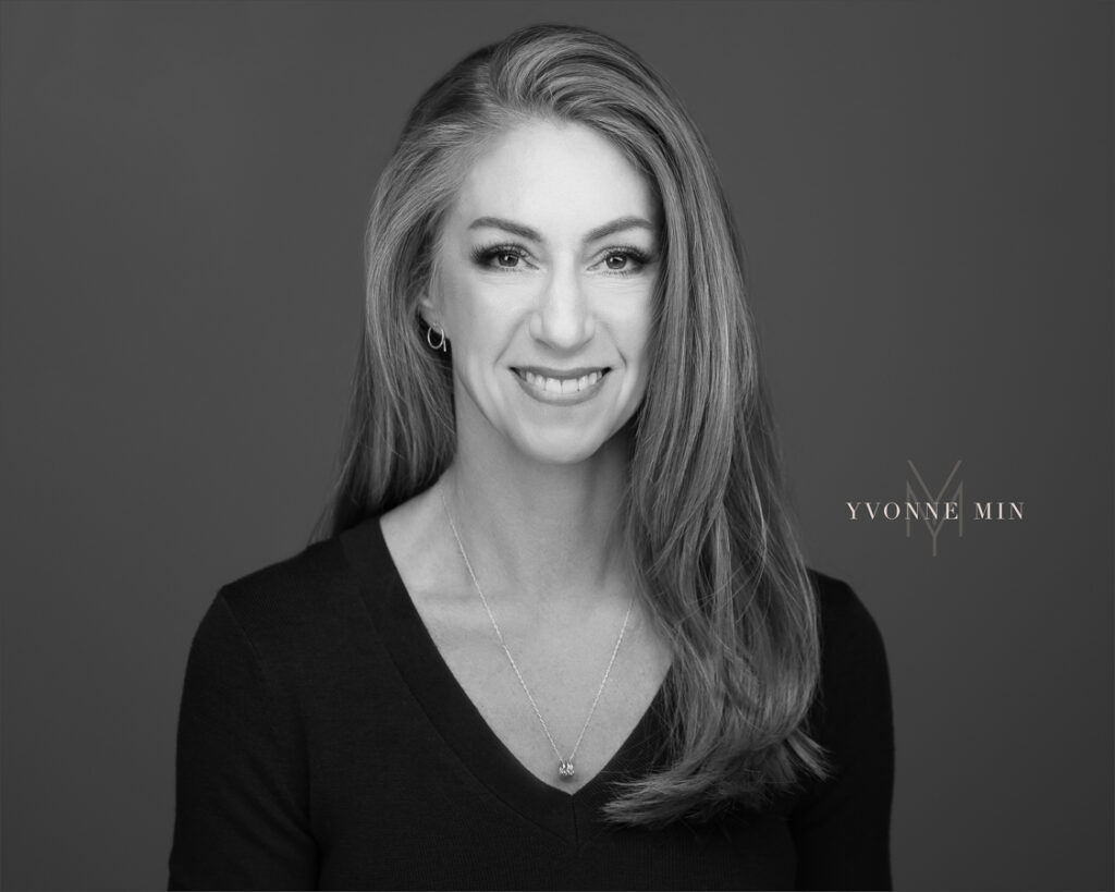 A black and white corporate headshot of a professional woman photographed on a dark gray backdrop at the studio of Yvonne Min Photography in Thornton, Colorado.