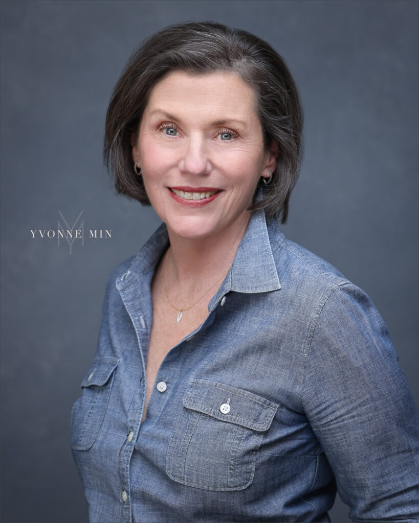 A headshot of a professional woman in a denim shirt photographed on a mottled dark gray backdrop in the studio of Yvonne Min Photography in Thornton, Colorado.