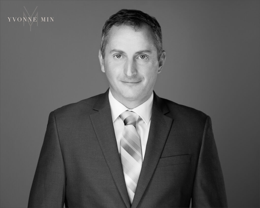 A new black and white headshot of a business professional in suit and tie on a dark gray backdrop photographed in the studio of Yvonne Min Photography in Thornton, Colorado.