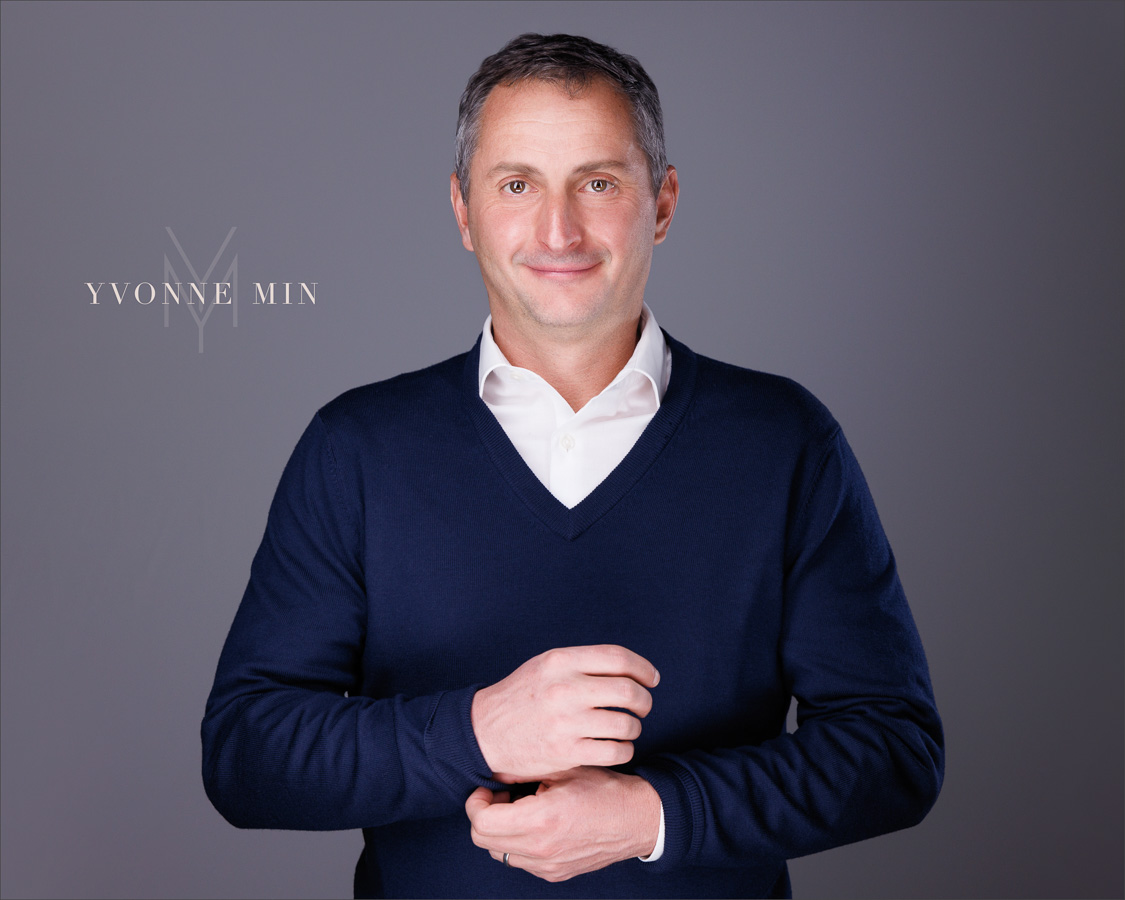 A new headshot of a business professional in a blue sweater on a dark gray backdrop photographed in the studio of Yvonne Min Photography in Thornton, Colorado.